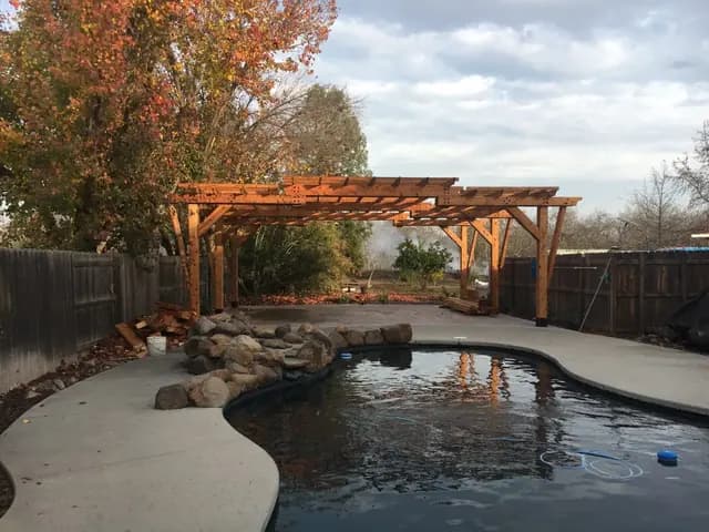 A cedar pergola with a stepped double canopy standing over a bare concrete pad beside a swimming pool in autumn, timber offcuts still stacked underneath.