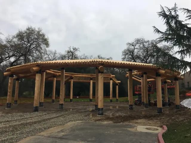 A large circular shade structure of peeled round log posts carrying a ring of log beams and close-spaced rafters, photographed on a wet day with a lift still on site.