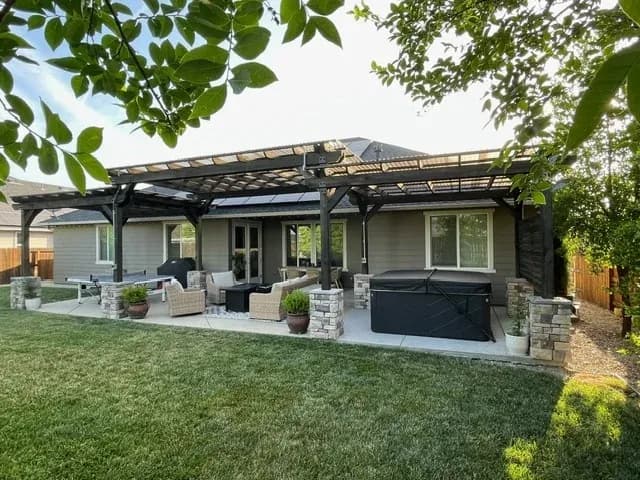 A long dark-stained pergola running the whole width of a modern house, carried on stone-clad piers over a patio with seating and a hot tub.