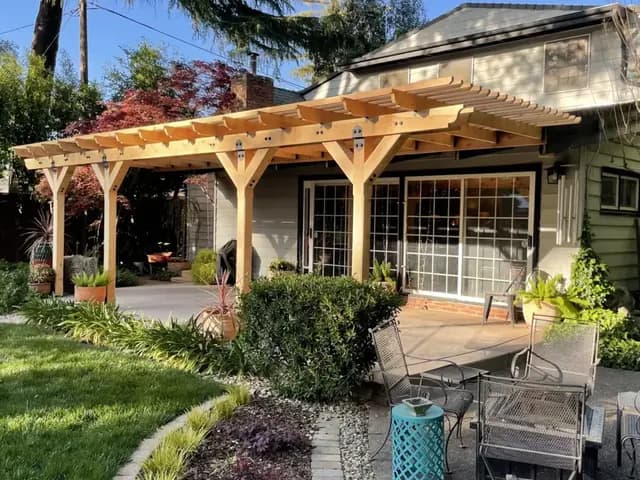 A pale cedar pergola attached along the back of a single storey house, its rafters running out over a patio, with planted borders and a lawn.