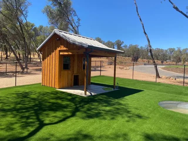 A small cedar-sided building with a standing seam roof and an open covered bay to one side, on artificial turf beside a stock tank.