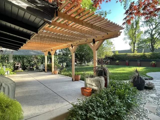 A cedar pergola on square posts running along a house wall beside a retractable awning, over a stamped concrete patio with potted plants.