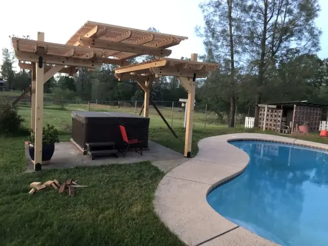 An unstained cedar pergola with two offset canopy tiers over a hot tub on a concrete pad, a kidney shaped swimming pool beside it.