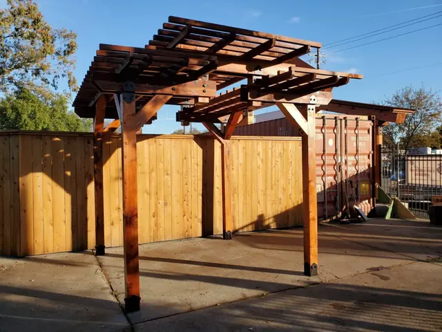 A stained cedar pergola with two stepped canopy tiers and steel plate connectors, standing complete on a concrete yard against a new board fence.