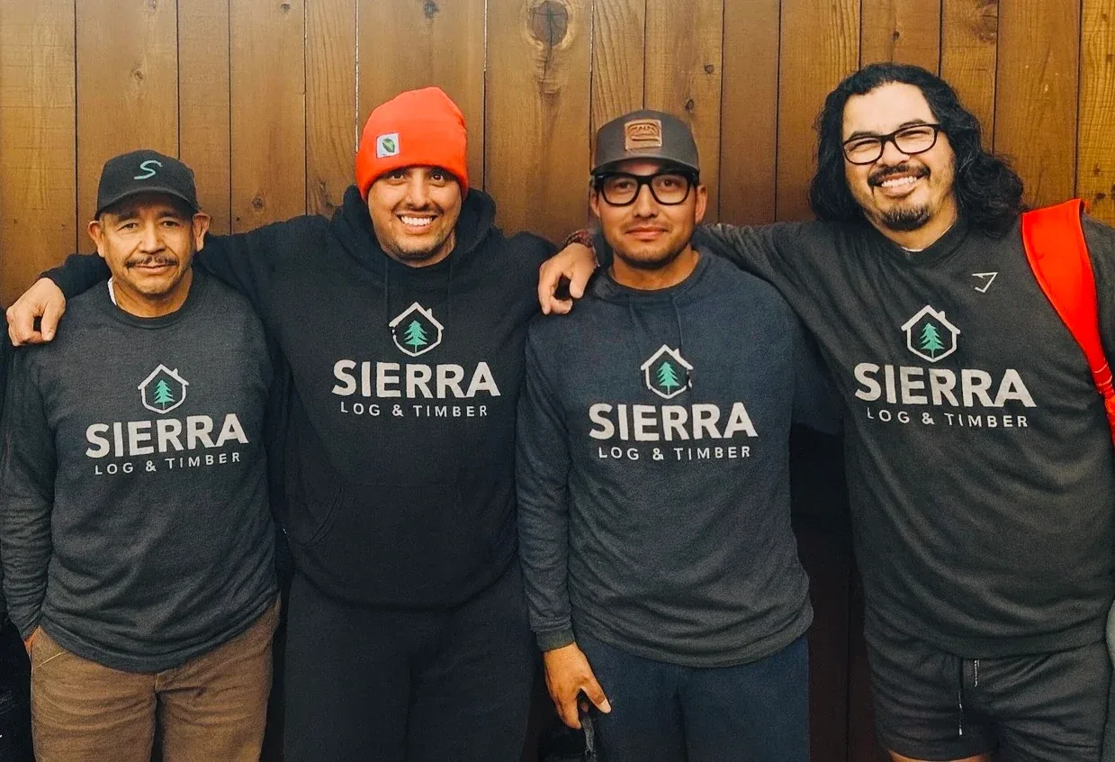 Four of the Sierra Log & Timber production crew in Sierra shirts, arms round each other in front of a cedar wall.