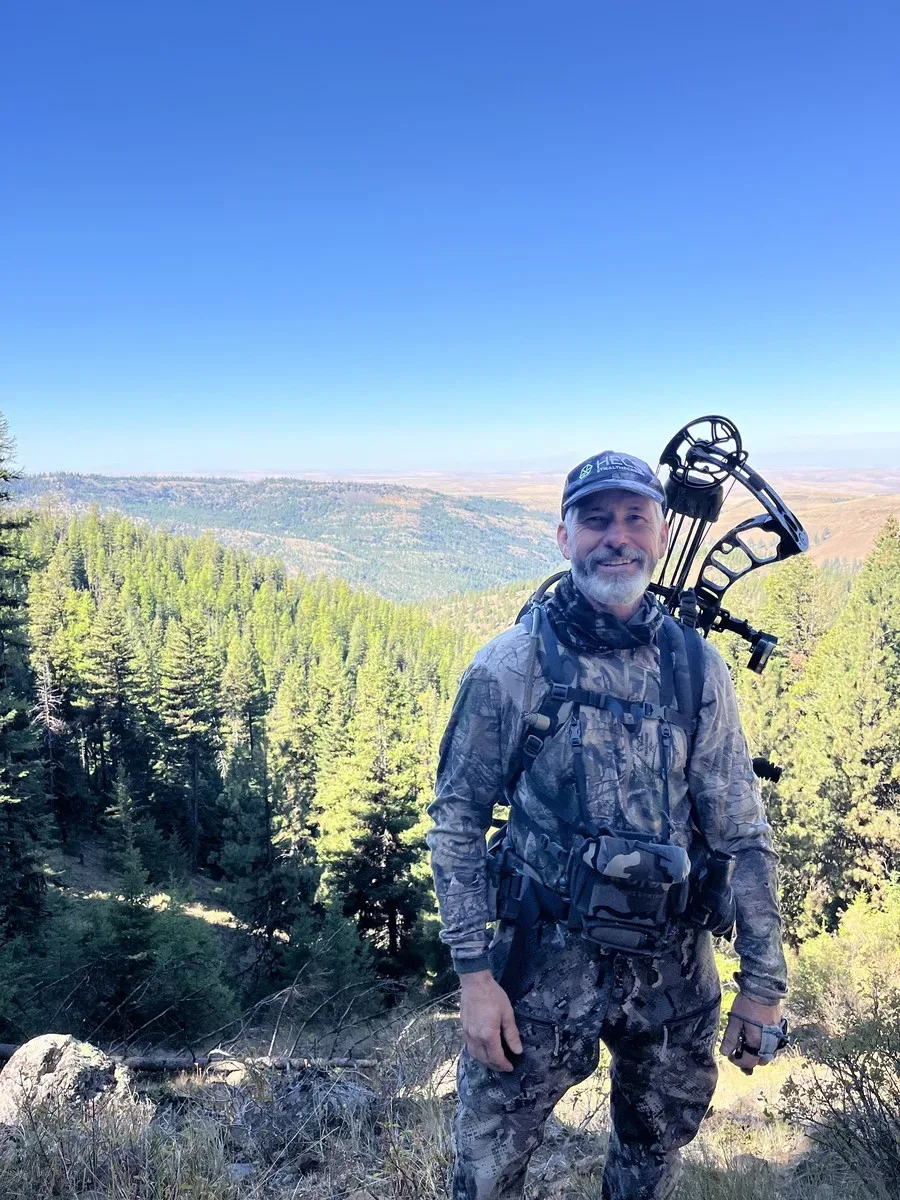 Hank Schaffeld in camouflage with a bow, on a ridge above a forested valley.