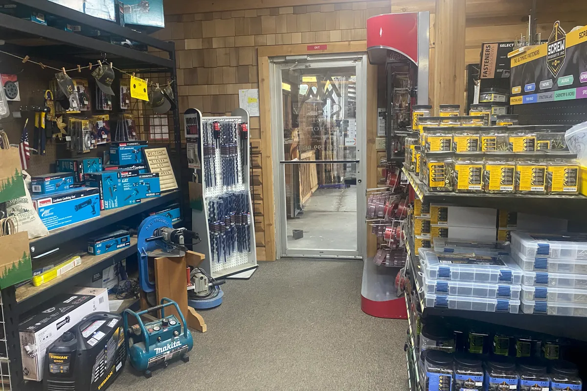 The aisle to the store's glass door: boxed Makita tools, hanging tools and a stand of long drill bits on the left, and on the right shelves of Screw Products fastener tubs and clear boxes of screws.