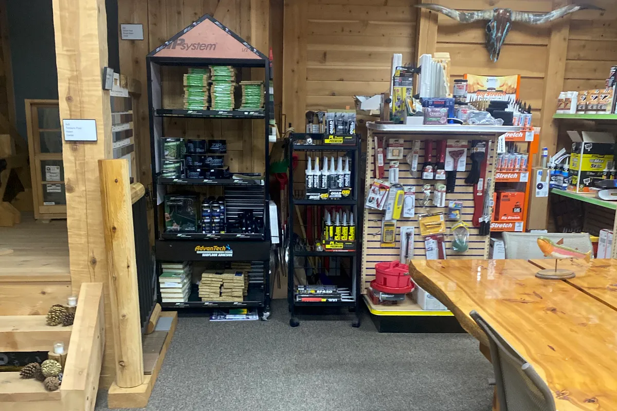 The back corner of the showroom: a ZIP System display with tape and AdvanTech subfloor adhesive, a cart of OSI caulk, a pegboard of hand tools and a caulk display under a longhorn skull, beside the end of the live-edge table and a log stair.