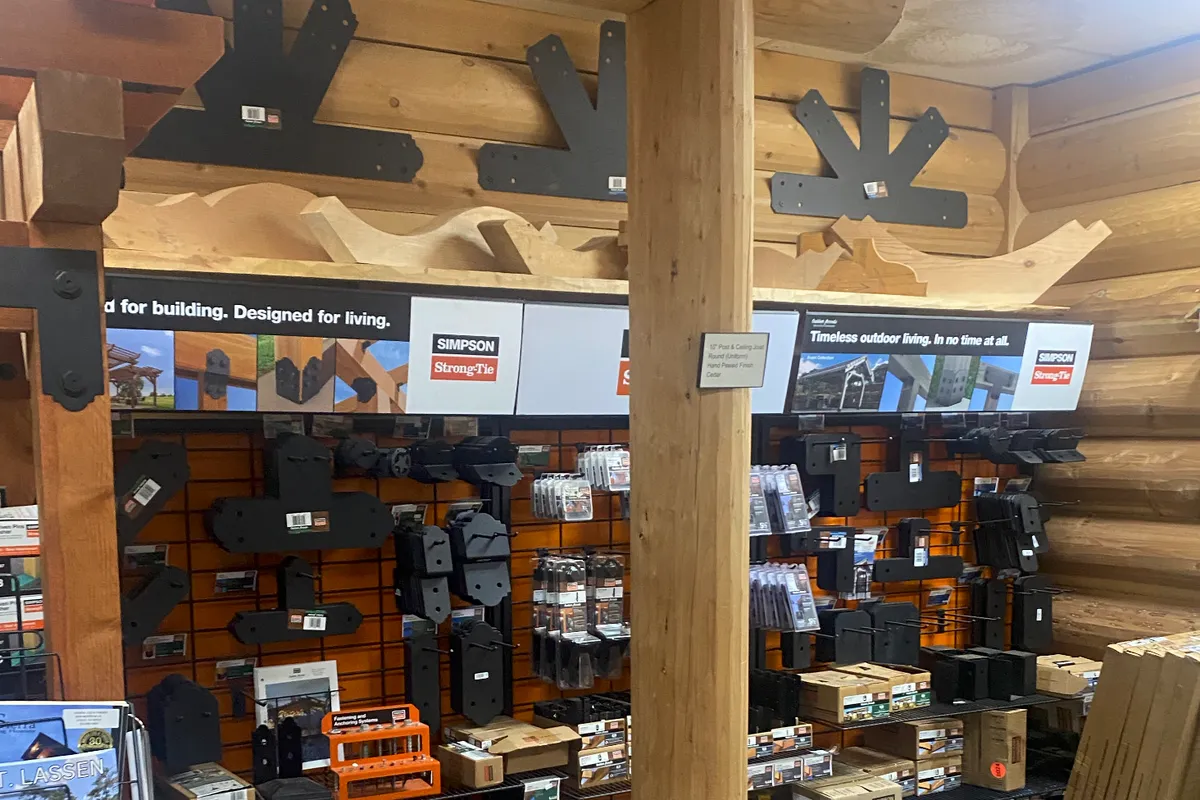 A wall of black Simpson Strong-Tie connectors and fastener packs on orange slatwall, under a ledge of curved log trim and large black post brackets mounted on the log wall above, with a round log post standing in front.