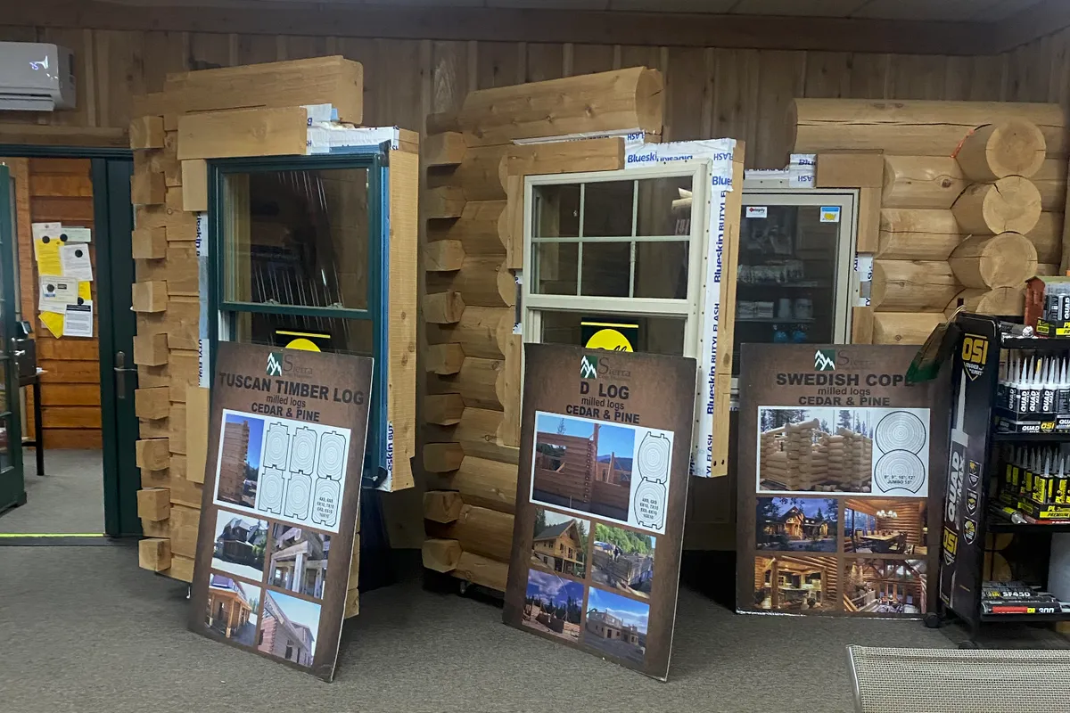 Three log wall corner samples in the showroom, each stacked around a window set in Blueskin flashing: squared timber logs, D-logs and round logs, with a board leaning in front of each that reads Tuscan Timber Log, D Log and Swedish Cope, milled logs, cedar and pine.