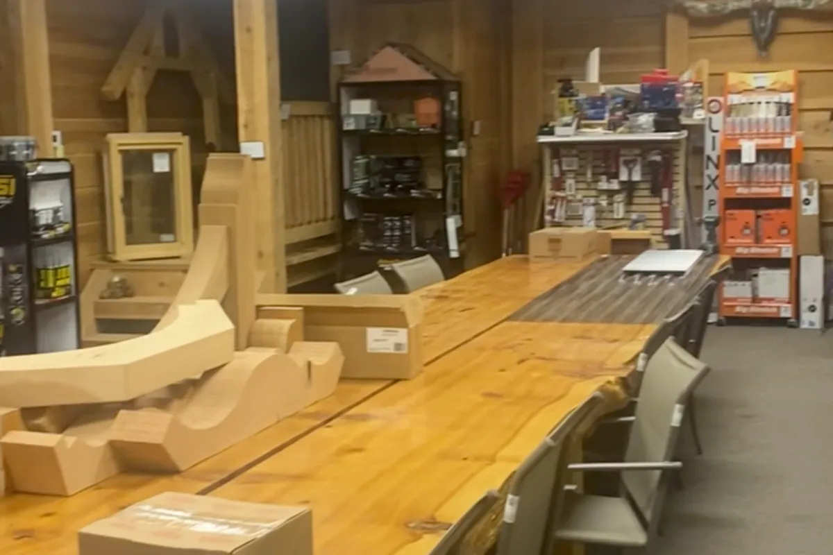 The showroom's long live-edge table, its glossy top running down the room past a row of chairs, with curved timber bracket pieces and a carton at the near end and tool and caulk displays along the back wall.