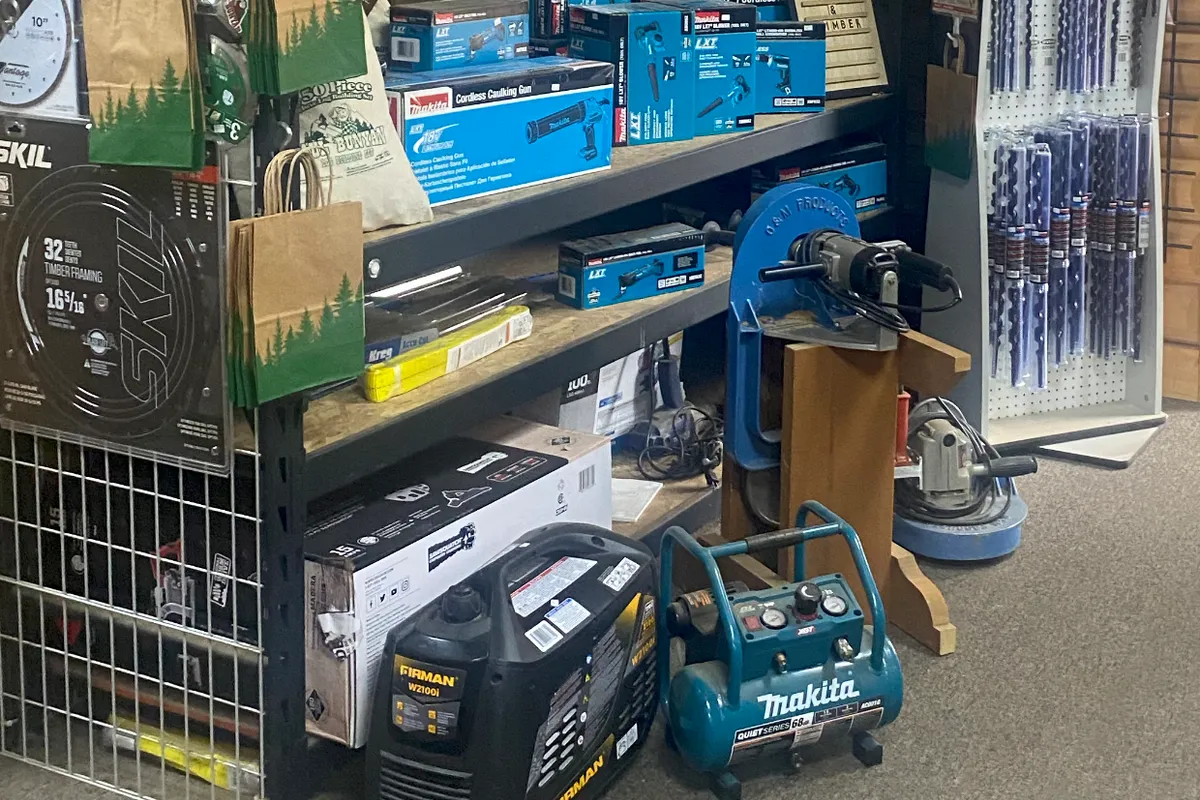 Shelving of boxed Makita LXT cordless tools, a cordless caulking gun among them, beside a Skil 16 5/16 inch timber framing blade, with a Firman generator and a Makita compressor on the floor, a heavy drill clamped in a blue stand, and a rack of long drill bits.