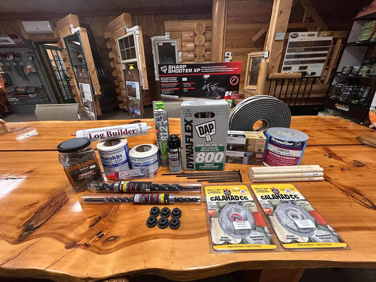 Log package sundries laid out on a live-edge table: a Sashco Log Builder tube, two rolls of Henry Blueskin ZeroFlash tape, a Lexel tube, a can of DAP 812 foam and a can of foam cleaner, a Sharp Shooter XP foam gun box, a box of DAP Dynaflex 800, a roll of gasket, two boxes of Simpson Strong-Tie screws, a roll of HydroGap SA, a bundle of wood dowels, a jar of Pico screws, loose long screws, two Wood Owl nail chipper bits, six hex nuts and two Galahad CG grinding discs.