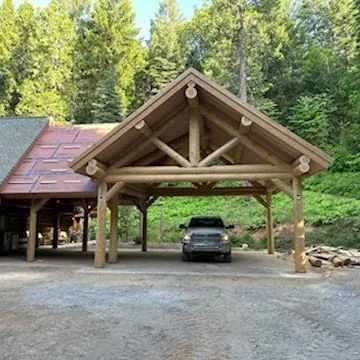 A finished log carport at the edge of a forest: peeled round log posts carrying a log truss and purlins under a gable roof, a pickup truck parked beneath it on a gravel drive.