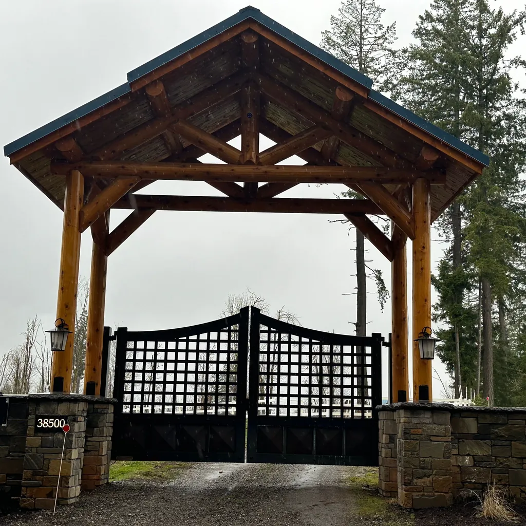A gated drive entrance: round log posts standing on stone pillars carry a timber truss and a small gable roof over black lattice gates, with tall firs behind.