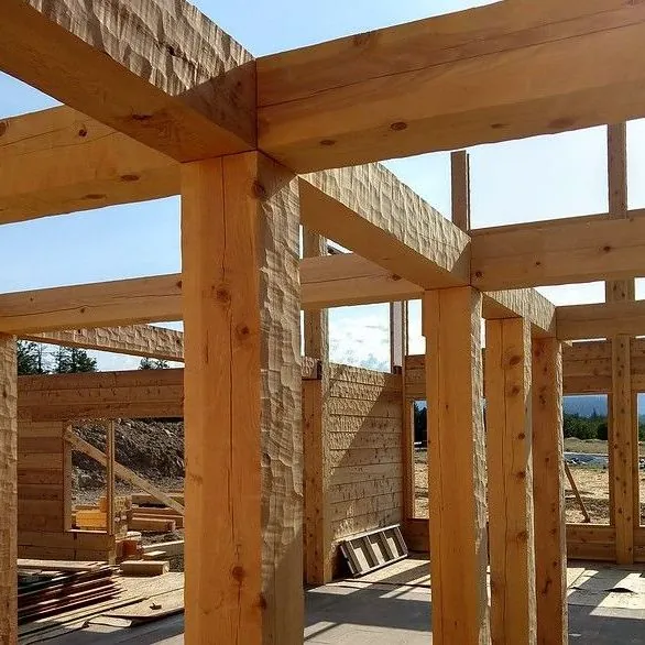 Heavy hand hewn timber girders resting on square timber posts in an open frame, their faces scalloped by the hewing tool, with a timber log wall and bright sky beyond.