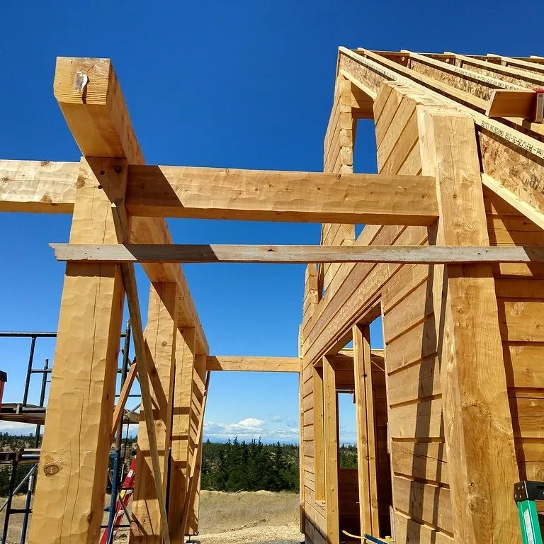 A hand hewn timber frame against a blue sky: square timber posts carrying a timber header across them, beside a wall of squared timber logs going up.
