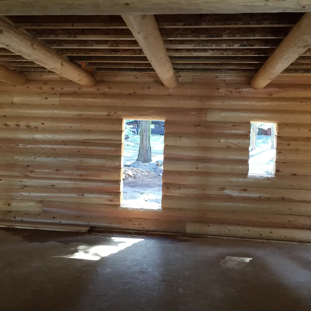 Inside a round log shell: peeled log floor joists span the room overhead with boards laid across them, above a wall of stacked logs with two window openings and a bare concrete floor.