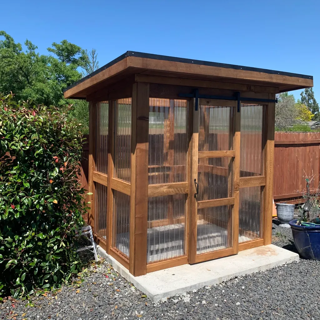 A small cedar greenhouse on a concrete pad: stained timber posts and rails frame clear ribbed panels on every side, with a sliding door on a black barn-door track and a flat roof overhanging all round.