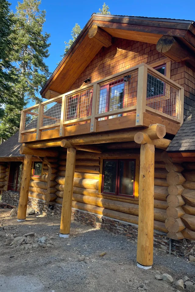 A round log home among pines with a new deck across its upper storey: tall natural log posts, peeled and tapering, stand on the gravel and carry log beams under the deck, whose timber railing is infilled with wire mesh, with cedar shingles in the gable above.
