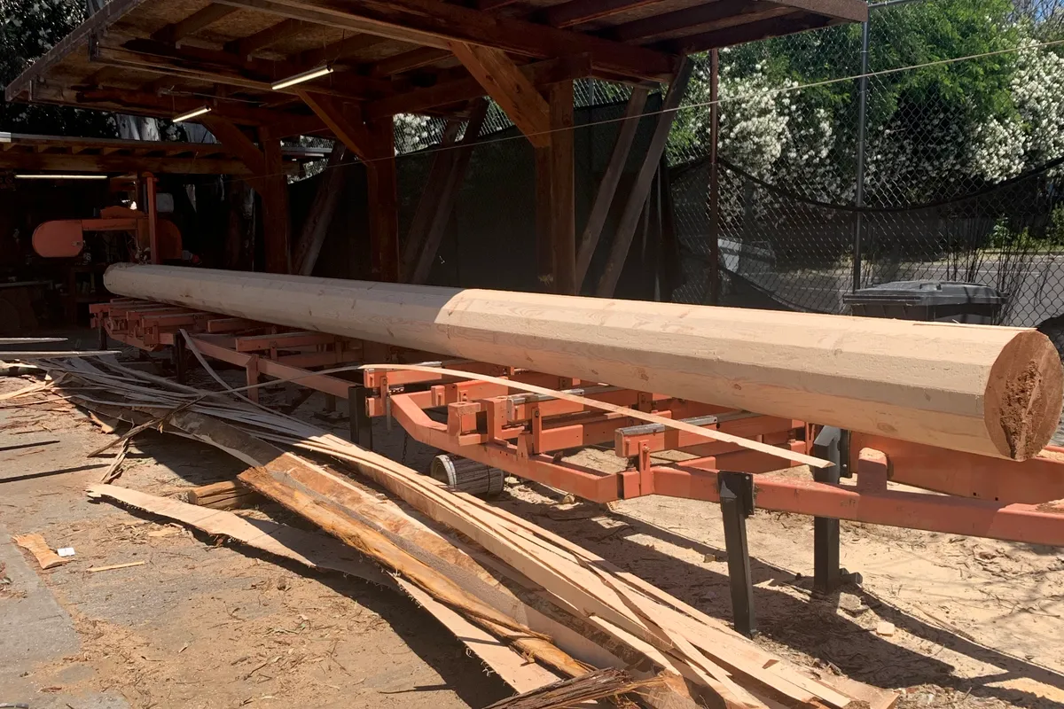 A long cedar log on a sawmill carriage under an open timber shed, already worked to one round diameter along its length, with the slabs taken off it piled on the ground below.