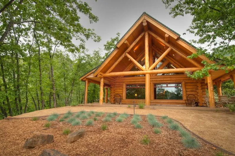 A log home's gable porch among oaks: natural peeled logs, each keeping its own taper and curve, form the posts, the truss and the beams over a deck, above a mulched bed of grasses and boulders.
