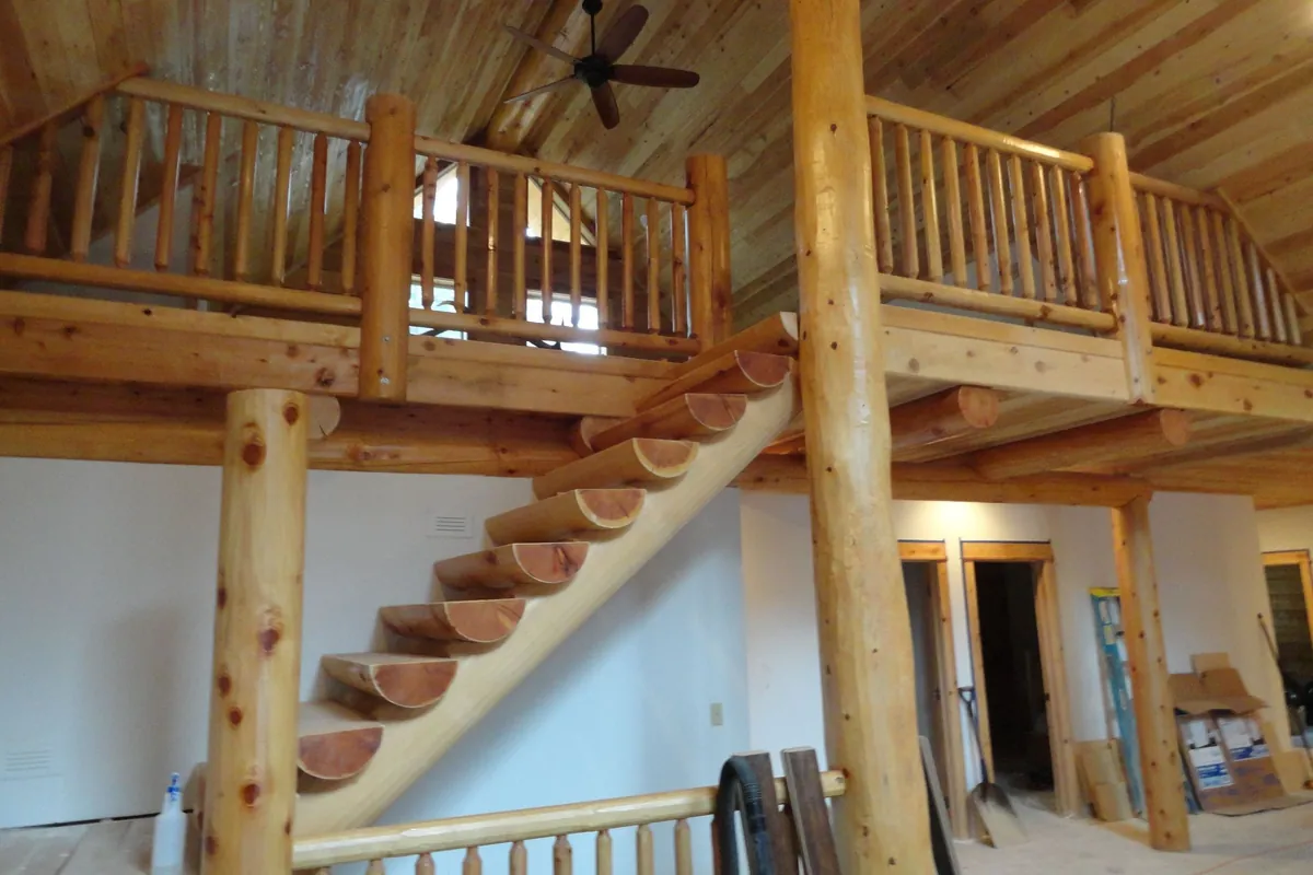 A loft and stair inside a log home before the finishes go in: uniform round logs, all one diameter, form the loft railing and the stair rails, beside tall natural log posts that keep their knots and taper, with half log treads climbing a log stringer.