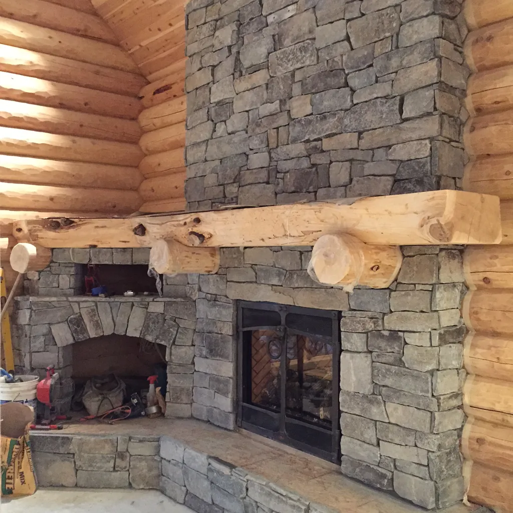A log mantel across a stacked stone fireplace in a round log room: a peeled half log resting on two short round log brackets, the stone chimney rising to the cedar ceiling above.