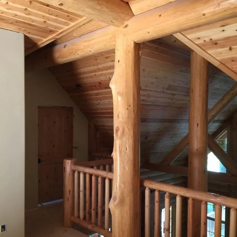 A natural log post with its taper and knots showing, standing at the top of a stair beside a round log railing and carrying a log beam under a tongue and groove cedar ceiling.