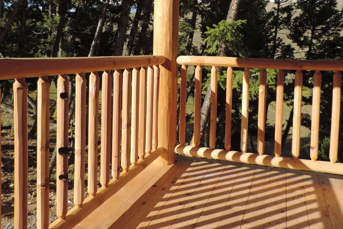 Close on a log railing at the corner of a deck in low sun: round log rails meet a log post, and the faceted round pickets between them, each tapered at its ends, throw striped shadows across the deck boards.