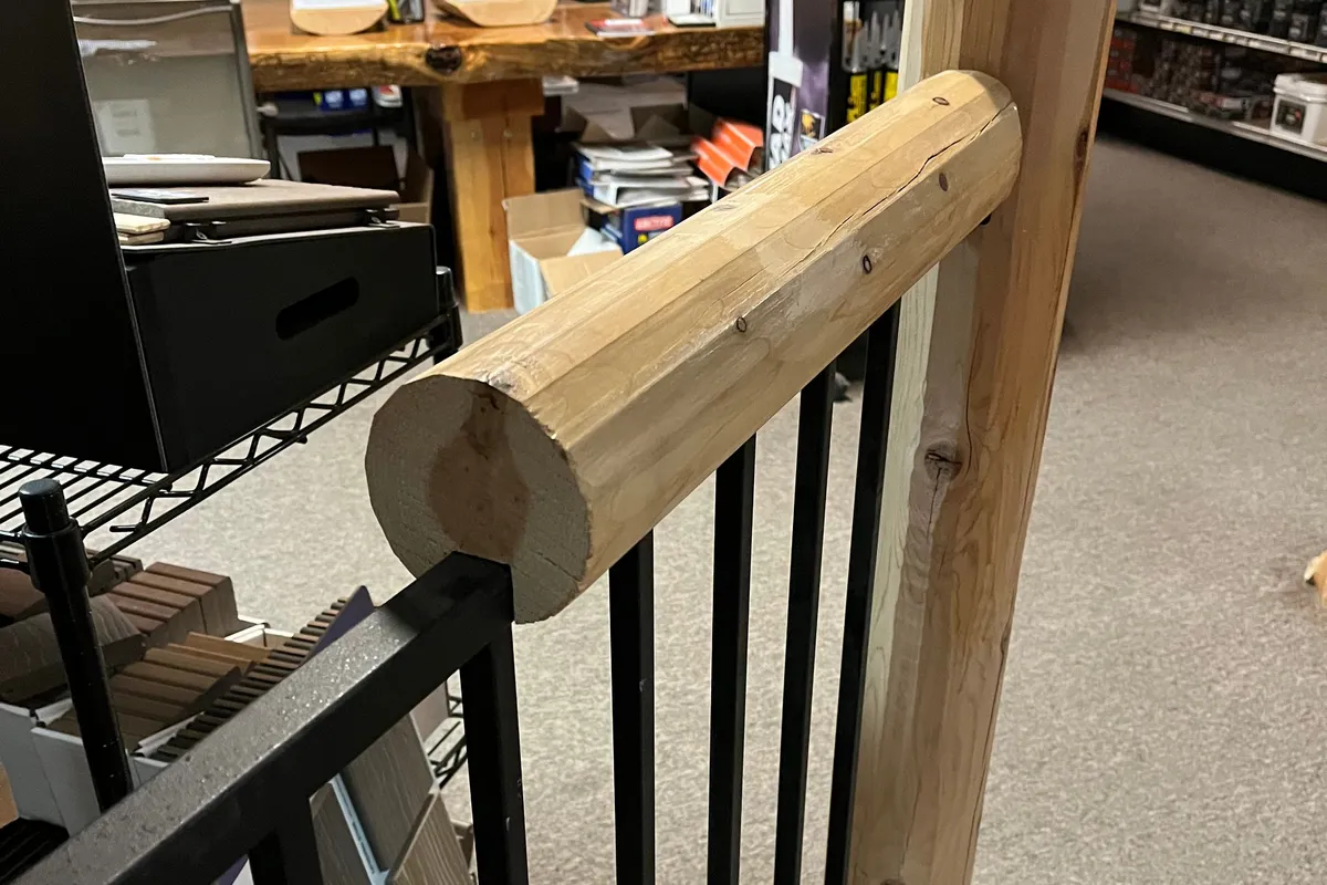 A round log rail cap, peeled and pale, running along a black steel baluster panel to a square timber post, set up in the showroom.