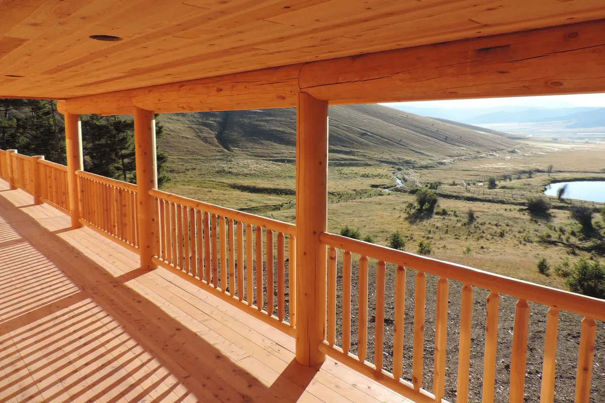 A long covered porch in low sun, looking out over a valley with a creek and a pond: round log railings with evenly spaced round pickets run between round log posts, under a log header and a pale board ceiling.