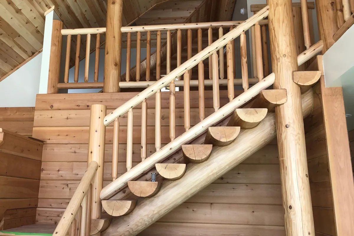A log stair inside a house, against walls of horizontal siding: half log treads, flat side up, climb a log horse, round pickets tapered at each end stand between a round log handrail and a lower rail, and a tall log post rises beside the stair, with a log railing round the loft above.