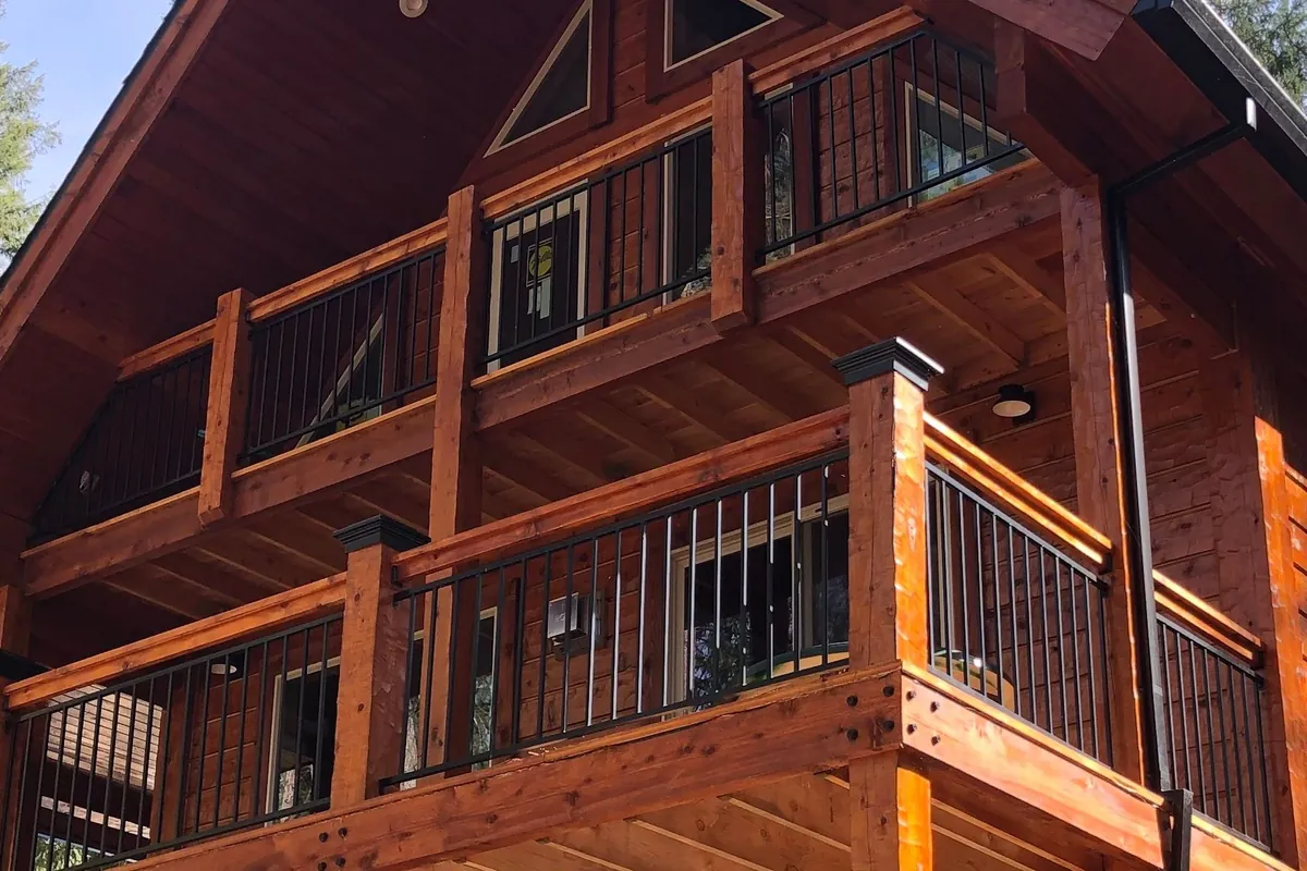 Two decks, one above the other, under the gable of a house, the wood stained a warm red-brown: square timber posts and timber top rails with black steel balusters between them, over heavy timber beams with black bolt heads at the joints.