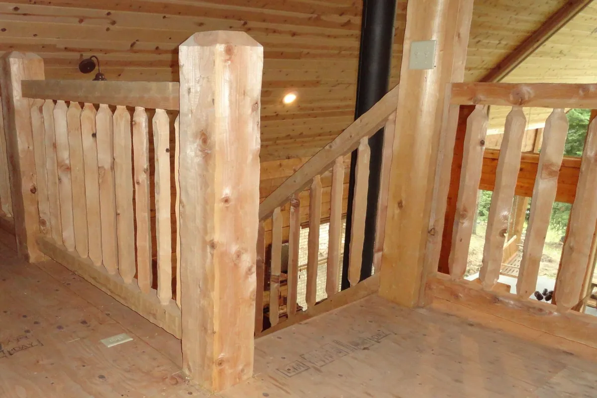 A timber railing round the top of a stair, over a plywood subfloor: square timber posts with chamfered tops, a timber top rail over flat timber pickets cut to a point at each end, and the stair rail running down beside a black pipe.
