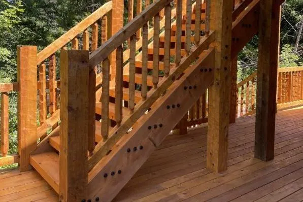 A timber stair rising from a deck among trees: square timber posts, timber handrails over shaped timber pickets, open treads between timber horses, and rows of three black fastener heads down the horse.