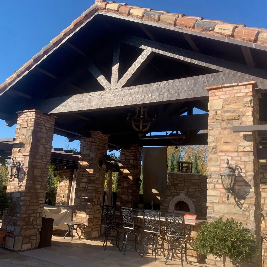 A large timber pavilion on stone columns: a dark stained truss and rafters under a clay tile roof, sheltering an outdoor dining table, wall lanterns and a stone pizza oven.