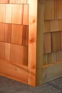 The same cedar shingles laid in even courses with a flush butt, meeting a rough sawn timber corner board at the edge of the wall.