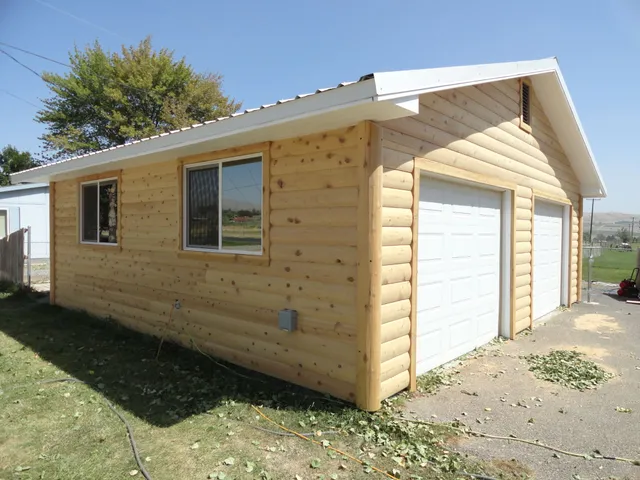 A detached two-car garage sided in unstained 2×8 log siding: the rounded courses run level along the side wall, around two windows, and up the gable end above two white garage doors.