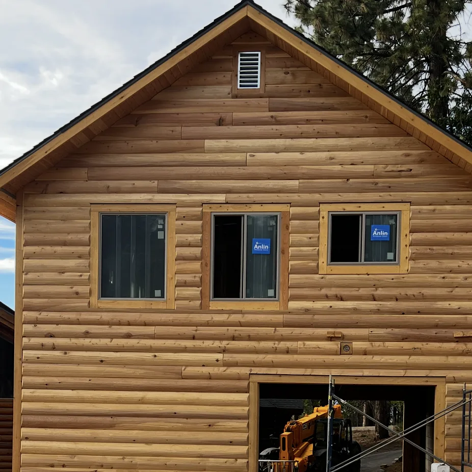 A new two-storey building clad to its gable in 3×10 log siding: long rounded courses with their joints staggered, three windows still wearing their labels, a louvred vent near the peak and a flat board trimming the corner.