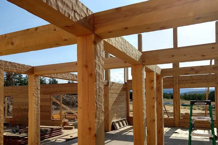 A timber frame going up on a building site: square posts and heavy beams, every face scalloped by the hewing tool, with a hewn timber wall rising behind them and a ladder among the posts.