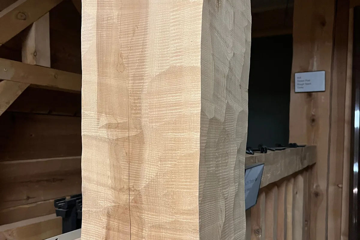 Close on a square hand hewn timber post in the showroom: shallow scallops left by the hewing tool run down its pale face, beside a wall of rough boards.