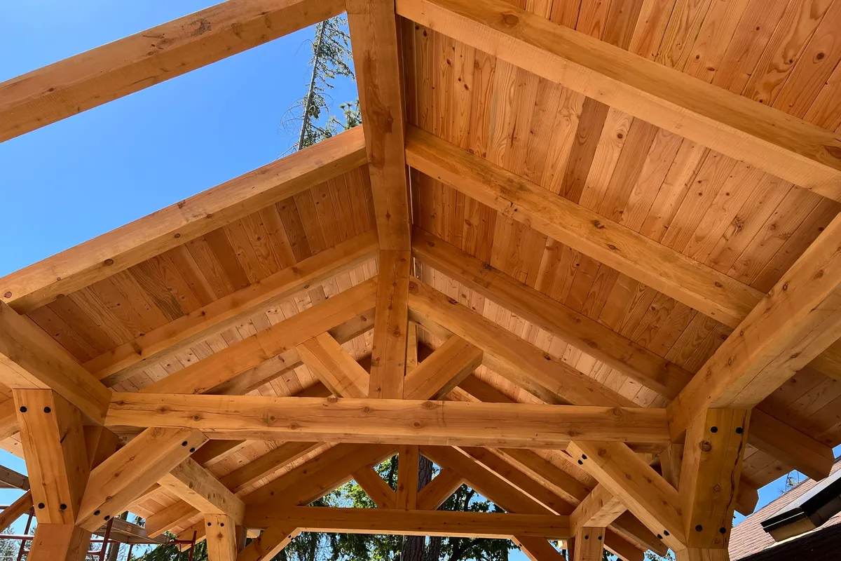 Looking up into a timber pavilion roof against a blue sky: rough sawn timber rafters, a king post truss and heavy beams meeting at the corners, under tongue and groove roof decking.