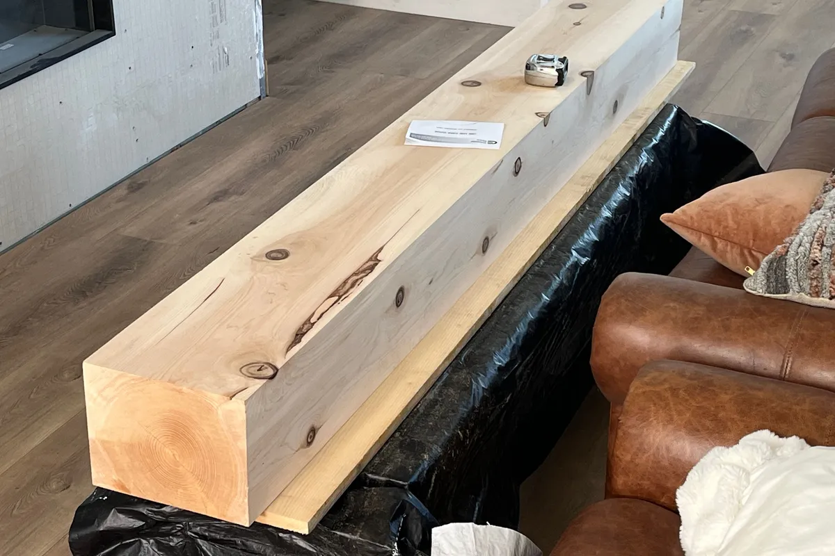 A long, smooth faced cedar timber laid on a sofa wrapped in plastic, ready to become a mantel: straight grain, a few dark knots and one streak of colour along its face.