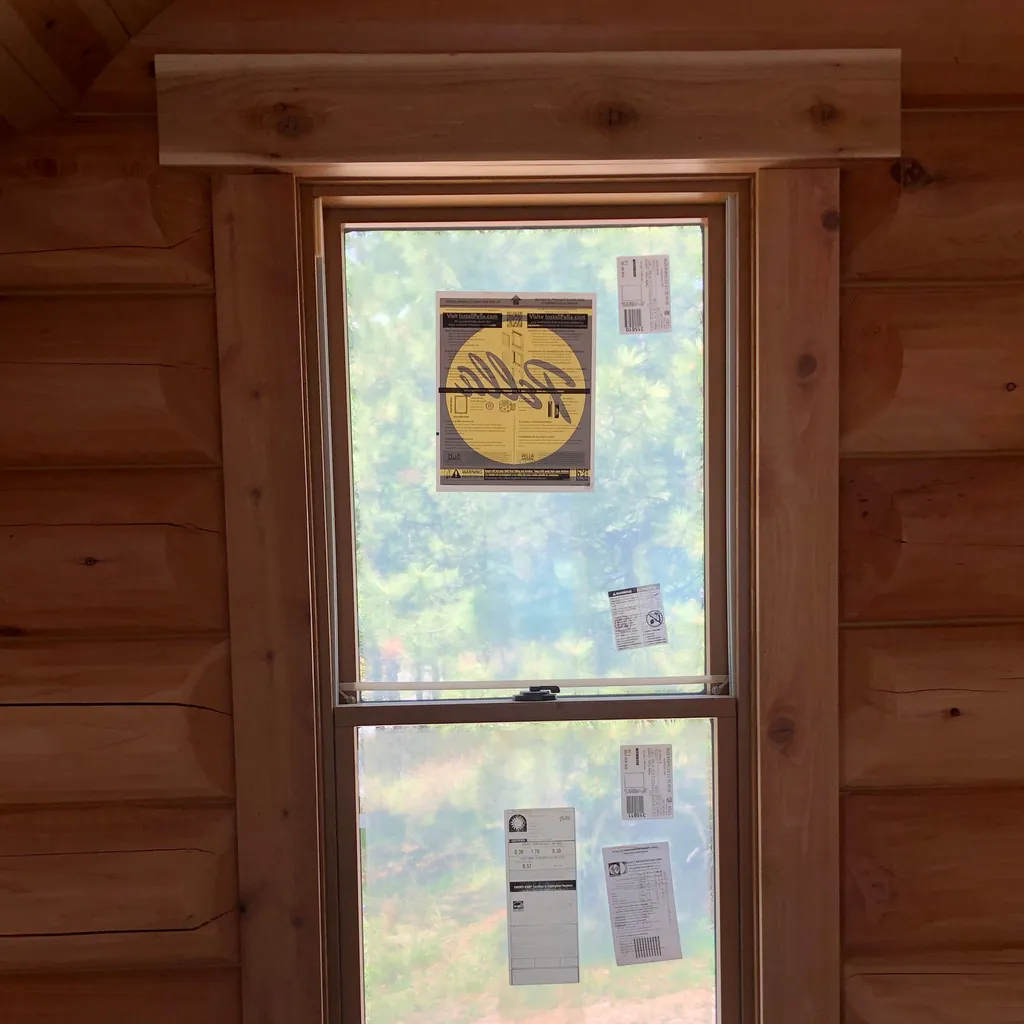 A new window set into a log wall and trimmed in flat cedar: a head casing with eared ends across the top and plain casings down both sides, the factory labels still on the glass.