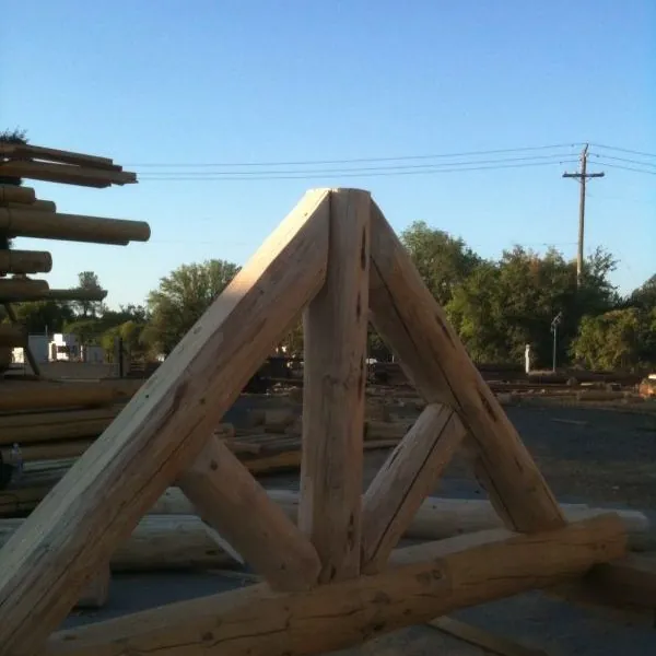 A peeled natural log truss standing on its own in a yard: two sloping rafters meeting at a king post, angled braces down to a long bottom chord, logs stacked at the left and power lines against the blue sky behind.