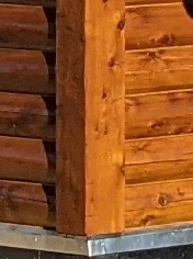 The corner of a D-log wall built against a square timber post: the rounded D-log courses run in from the left and stop against the post, flatter courses run off to the right, and the post stands on a metal sill flashing.