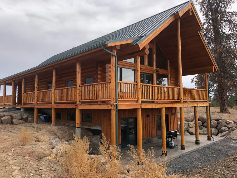 A round log home with a standing-seam metal roof and a deep covered deck, log ends crossing at every corner, on a dry hillside.