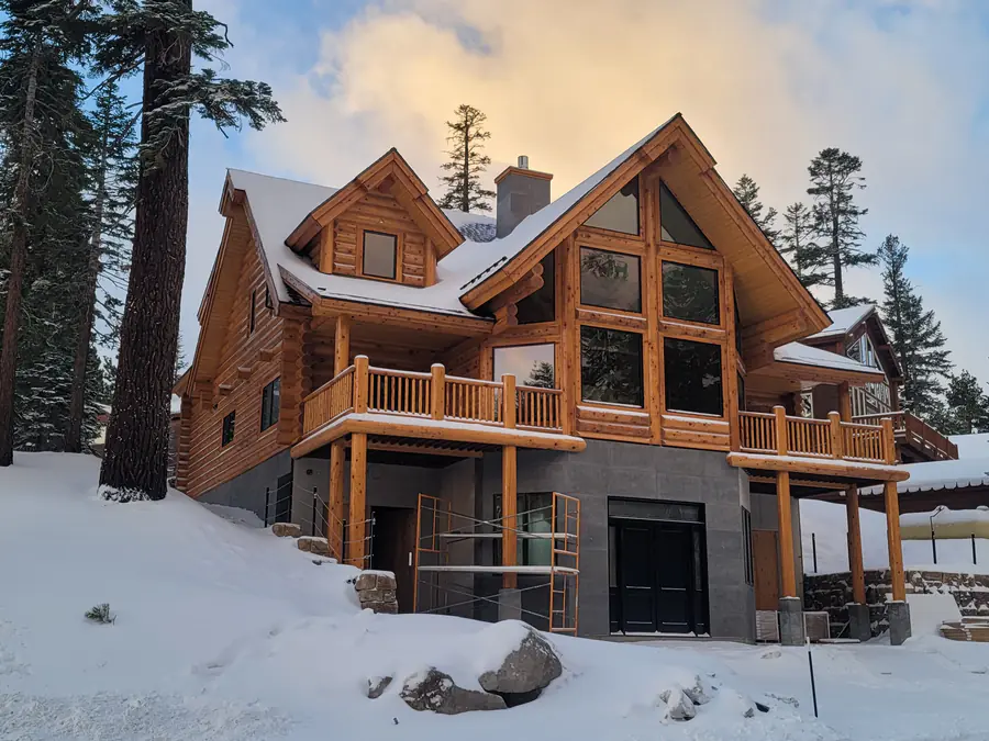 A round log home in snow, its prow gable glazed floor to roof and the log courses and crossed corners dusted white.