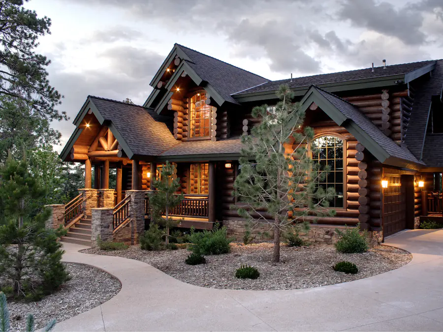 A finished round log home at dusk with stone piers, gable dormers and planted beds, the log corners stacked and notched.